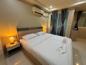 A hotel room with a bed, bedside lamp, air conditioning unit, curtains, and a small table with a kettle. - Lexis Residence Patong - Newly Renovated (Patong)