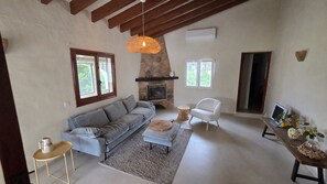 Un salon avec une cheminée en pierre, un canapé, un fauteuil blanc, une table basse ronde et une table d’appoint en bois sur laquelle se trouvent un ordinateur portable et des objets décoratifs.