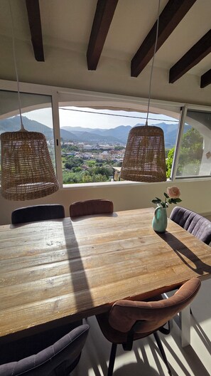 Un espace repas avec une grande table en bois, des suspensions en osier et une vue sur un paysage montagneux grâce à de grandes fenêtres.