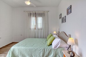 A bedroom with a bed, a nightstand, a lamp, a ceiling fan, and a window with sheer curtains.