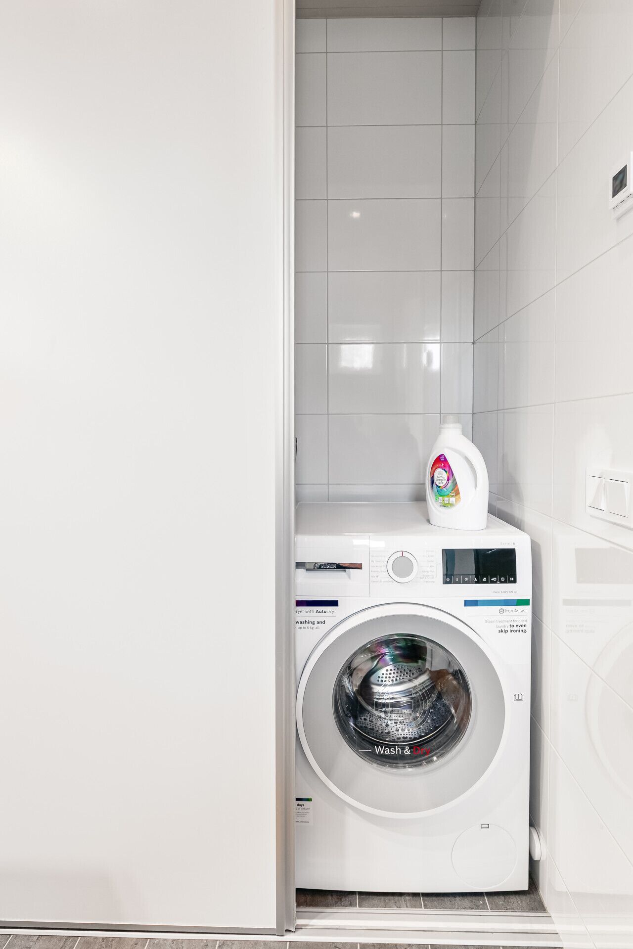 A white washing machine with a control panel, a detergent bottle, and a tiled wall.