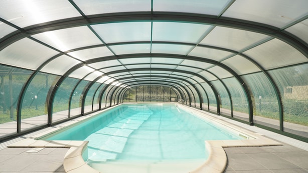 Ein Schwimmbad mit Glasdach, umgeben von einer gefliesten Terrasse.