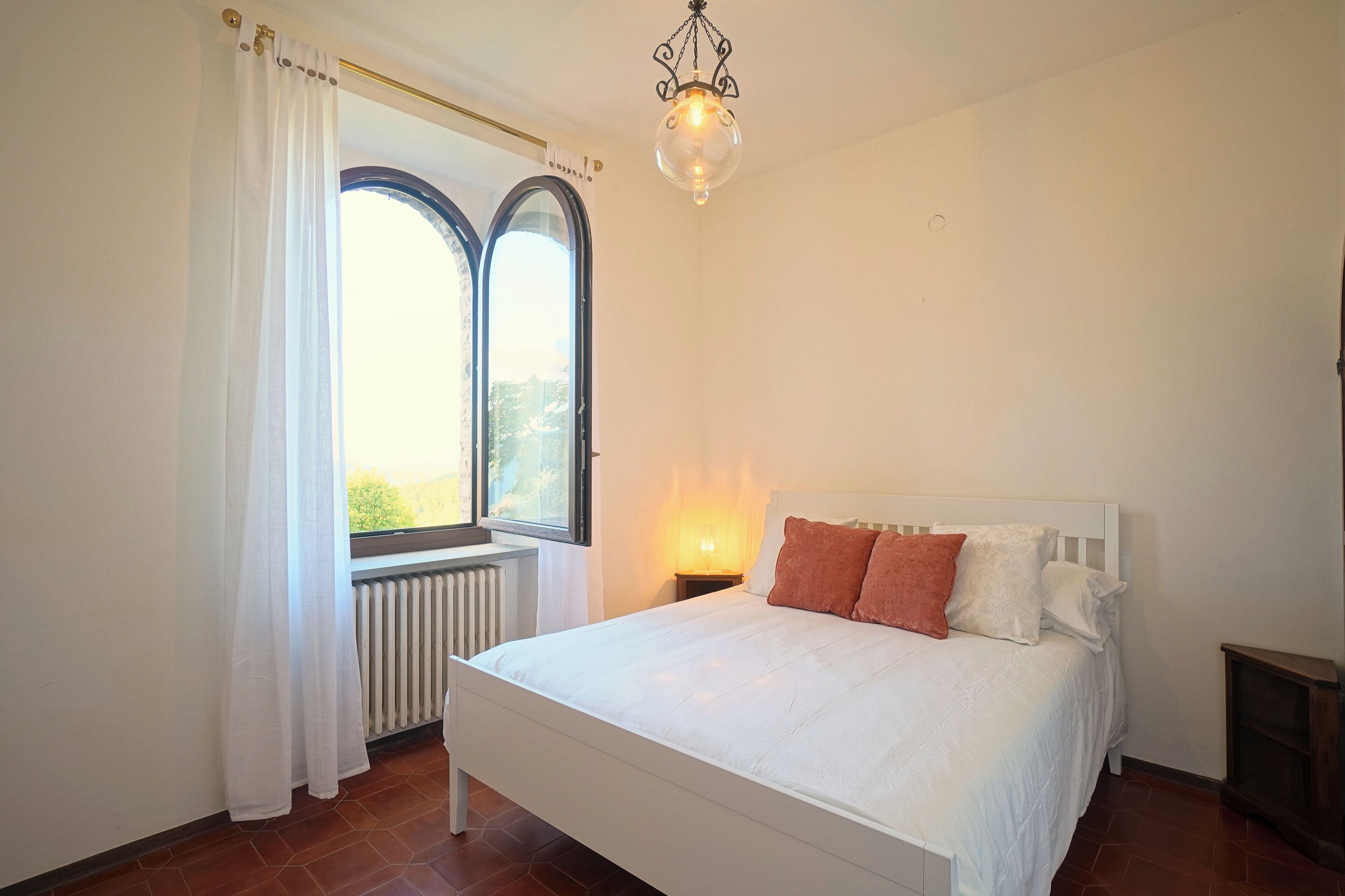 A bedroom with a bed, a window with curtains, a radiator, a nightstand with a lamp, and a chandelier.