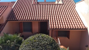 A residential building with terracotta tiled roofs, a small garden with a neatly trimmed bush, and a clear view of the sea in the background.