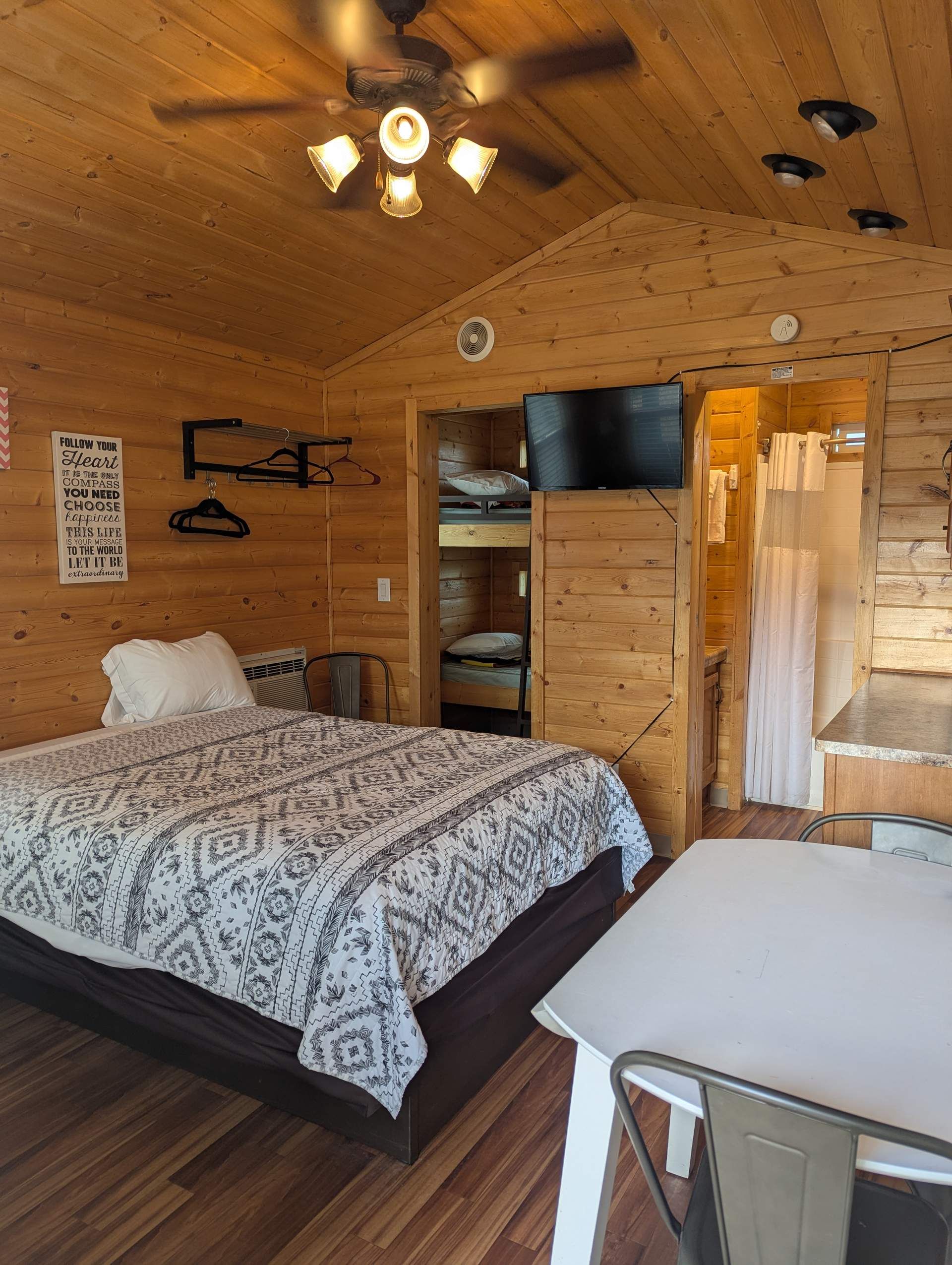 A wooden cabin room with a bed, a table, a TV, and a ceiling fan.
