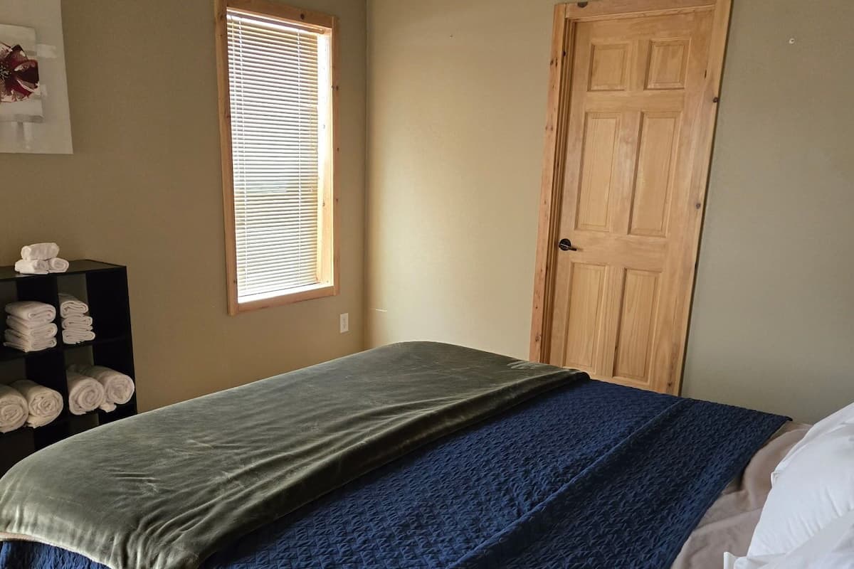 A bedroom with a bed, a wooden door, a window with blinds, and a shelf with towels.