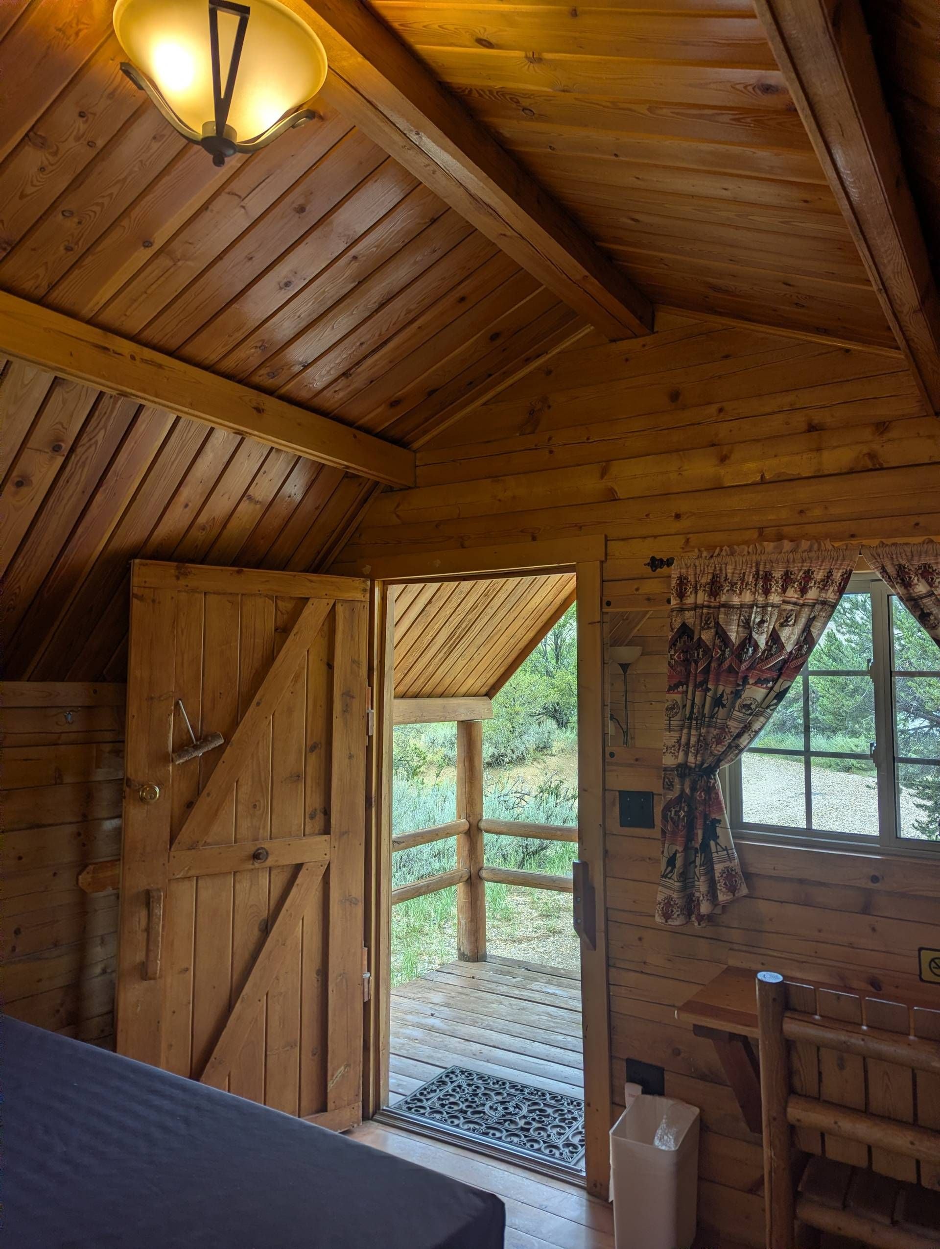 Interior de una cabaña de madera con cama, una puerta que da al exterior y una ventana con cortinas.