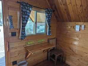 A cabin interior with a window, curtains, a bench, a small stool, and a trash bin.