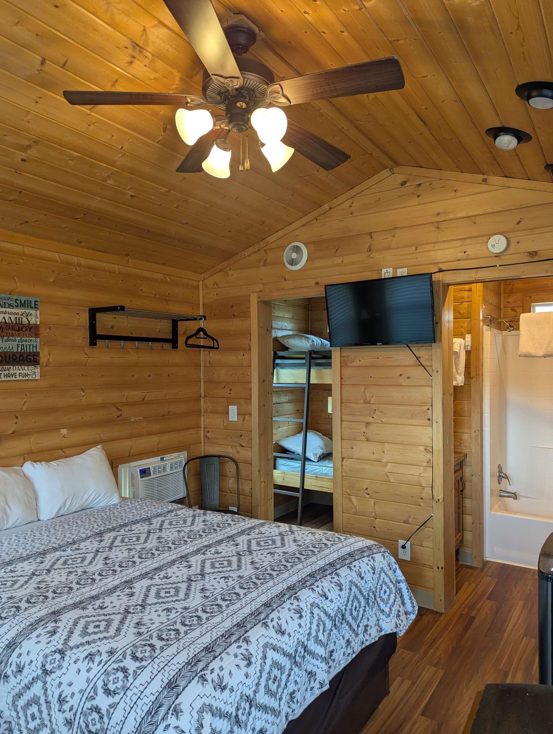 A wooden cabin room with a bed, a ceiling fan, a television, and a small bathroom.