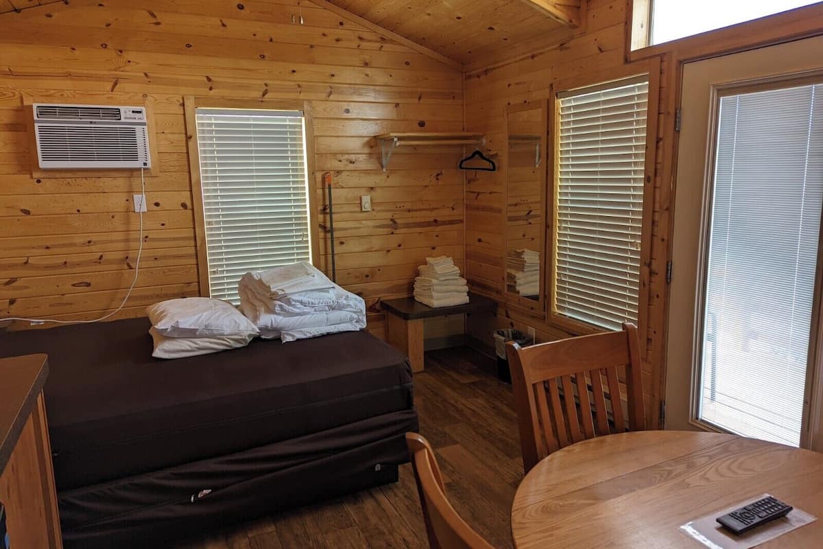 A wooden cabin room with a bed, a table, a chair, a ceiling fan, and a window with blinds.