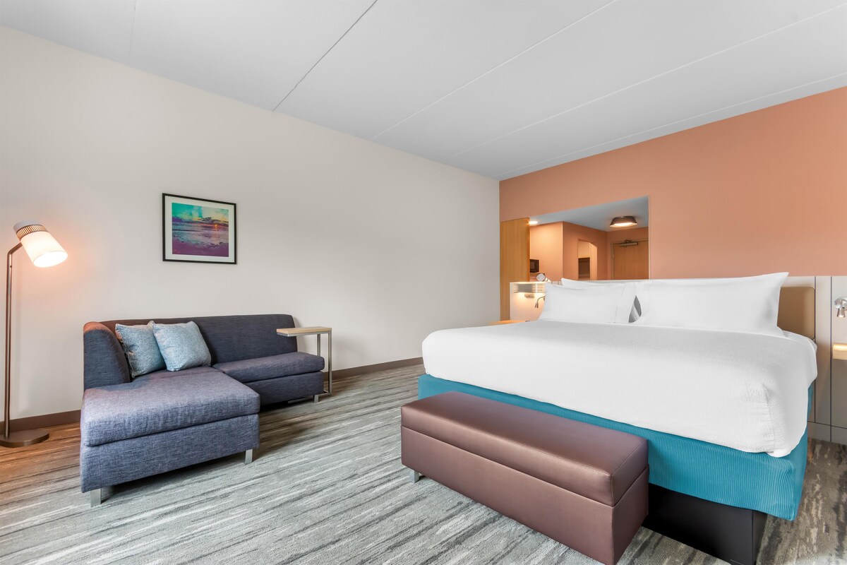 A hotel room with a bed, a sofa, a bench, a nightstand, a lamp, and a painting on the wall.