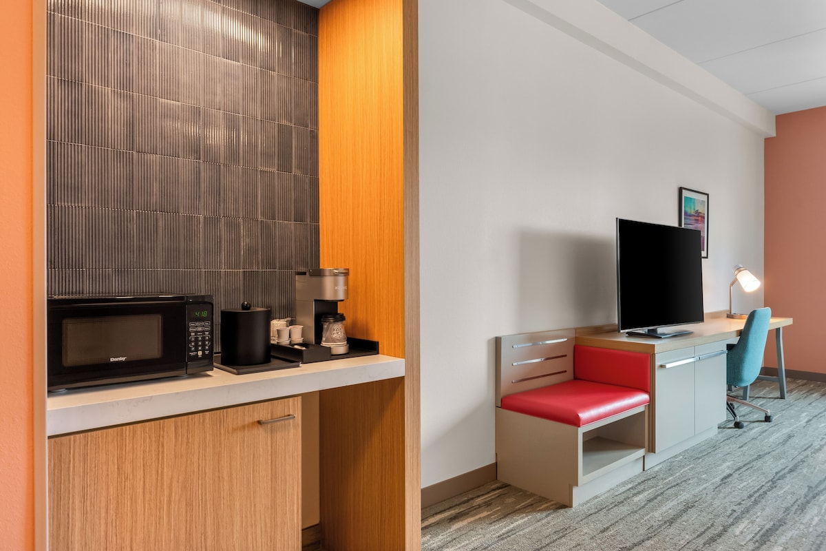 A hotel room with a kitchenette, a television, a desk with a chair, and a small window.