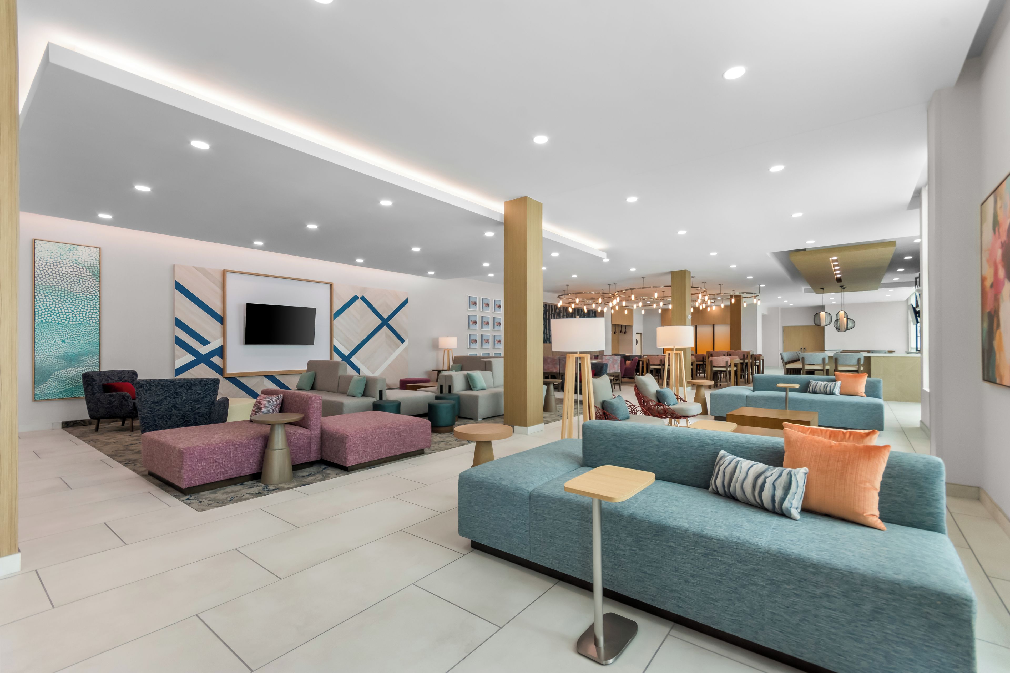 A modern hotel lobby with a mix of seating areas, a flat-screen TV mounted on the wall, and a dining area in the background.