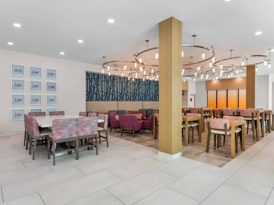 Uma área de refeições moderna com várias mesas e cadeiras, uma parede decorativa e um teto com luminárias circulares.