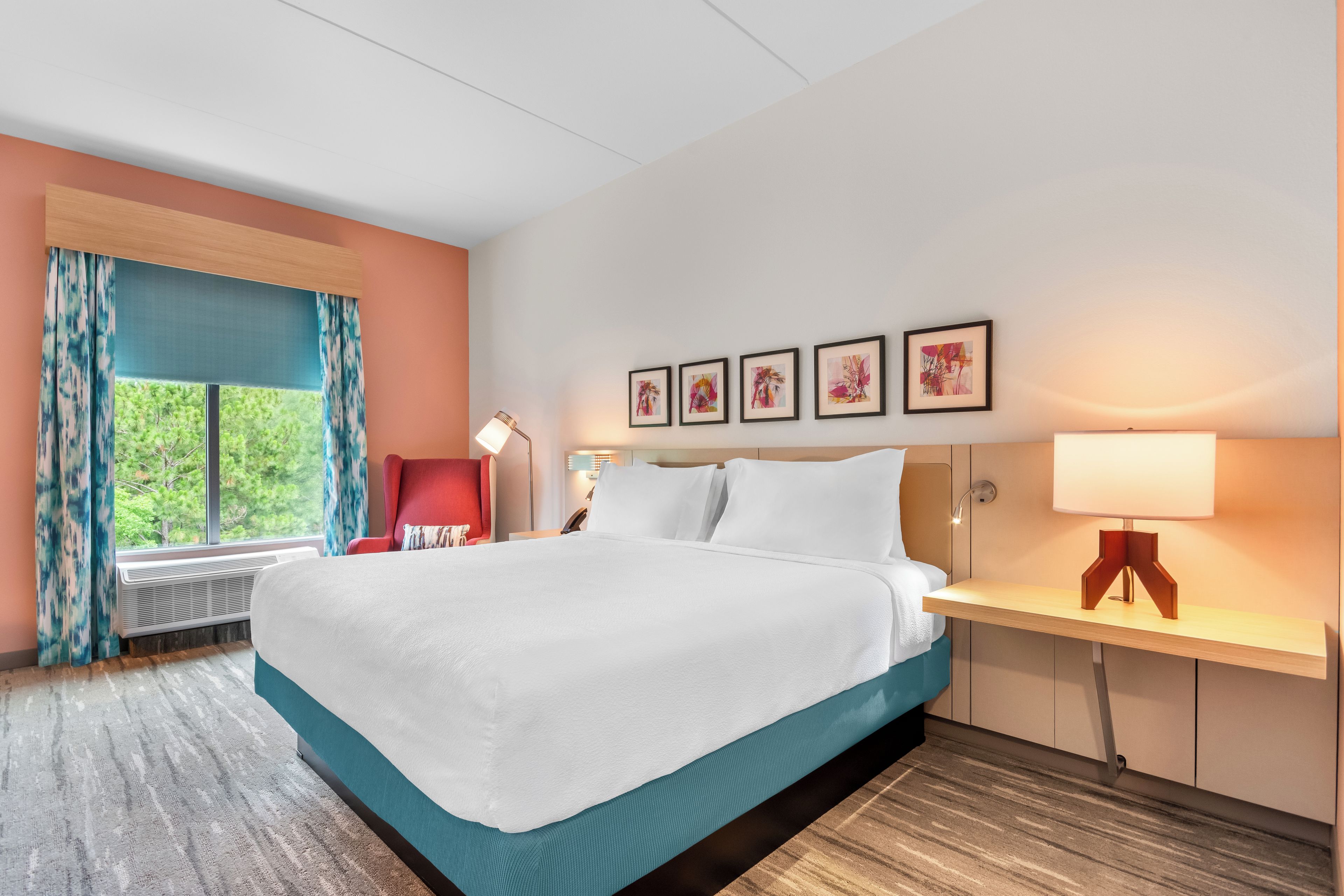 A hotel room with a bed, a desk, a chair, a window with curtains, and wall art.