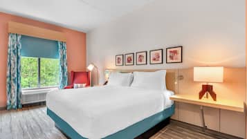 A hotel room with a bed, a desk, a chair, a window with curtains, and wall art.