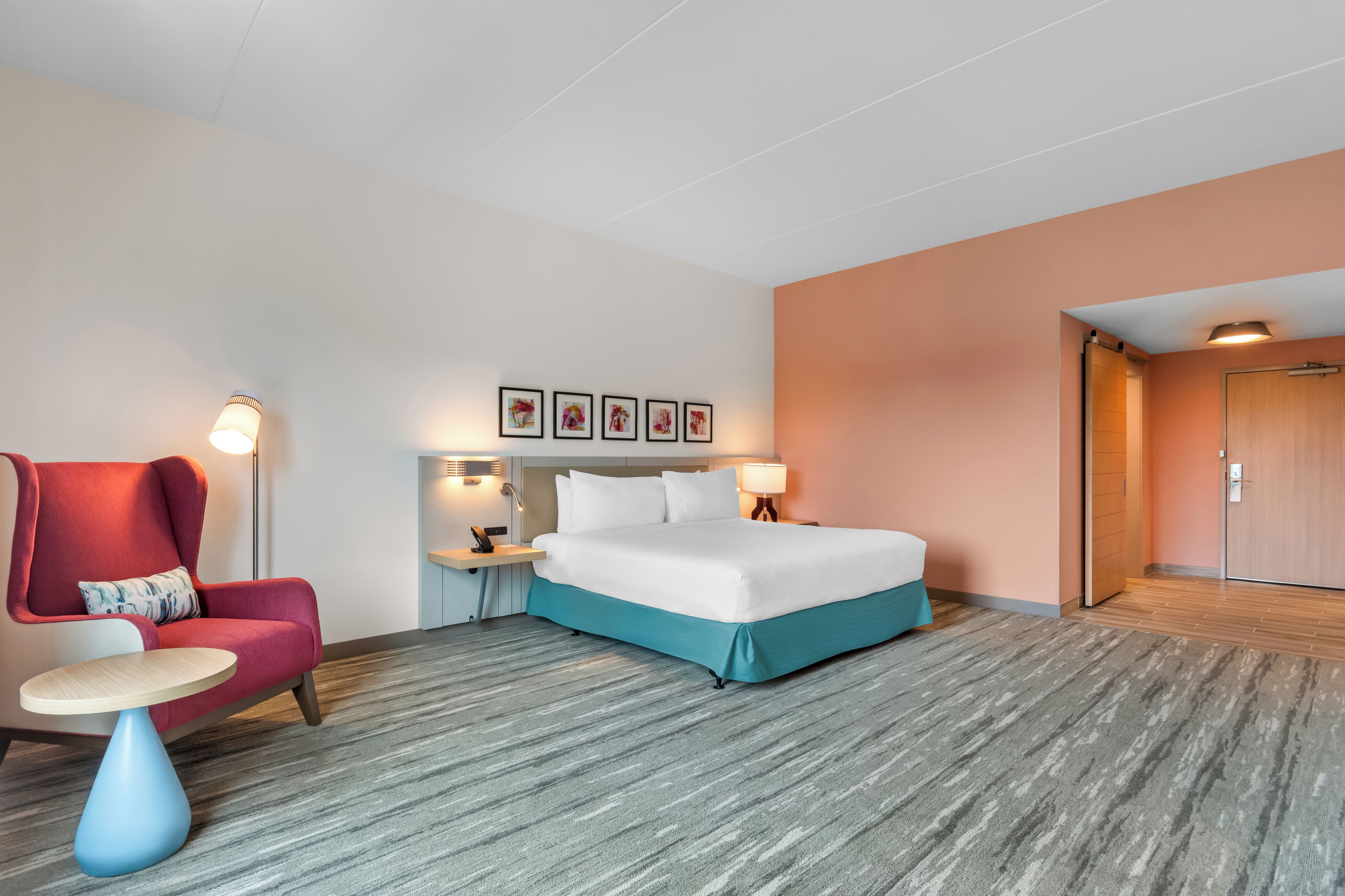 Une chambre d’hôtel avec un lit, une chaise, une petite table, une table de chevet et un mur orné de tableaux.