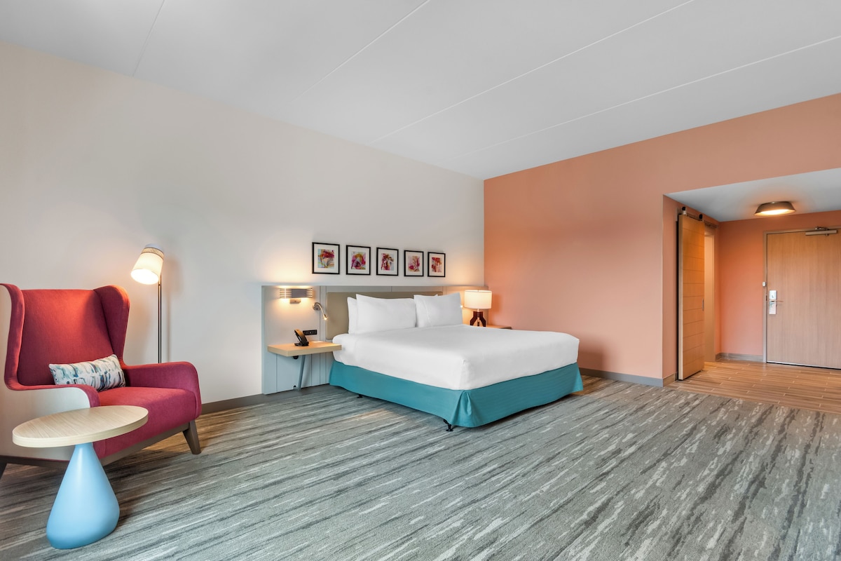 A hotel room with a bed, a chair, a small table, a nightstand, and a wall with framed pictures.