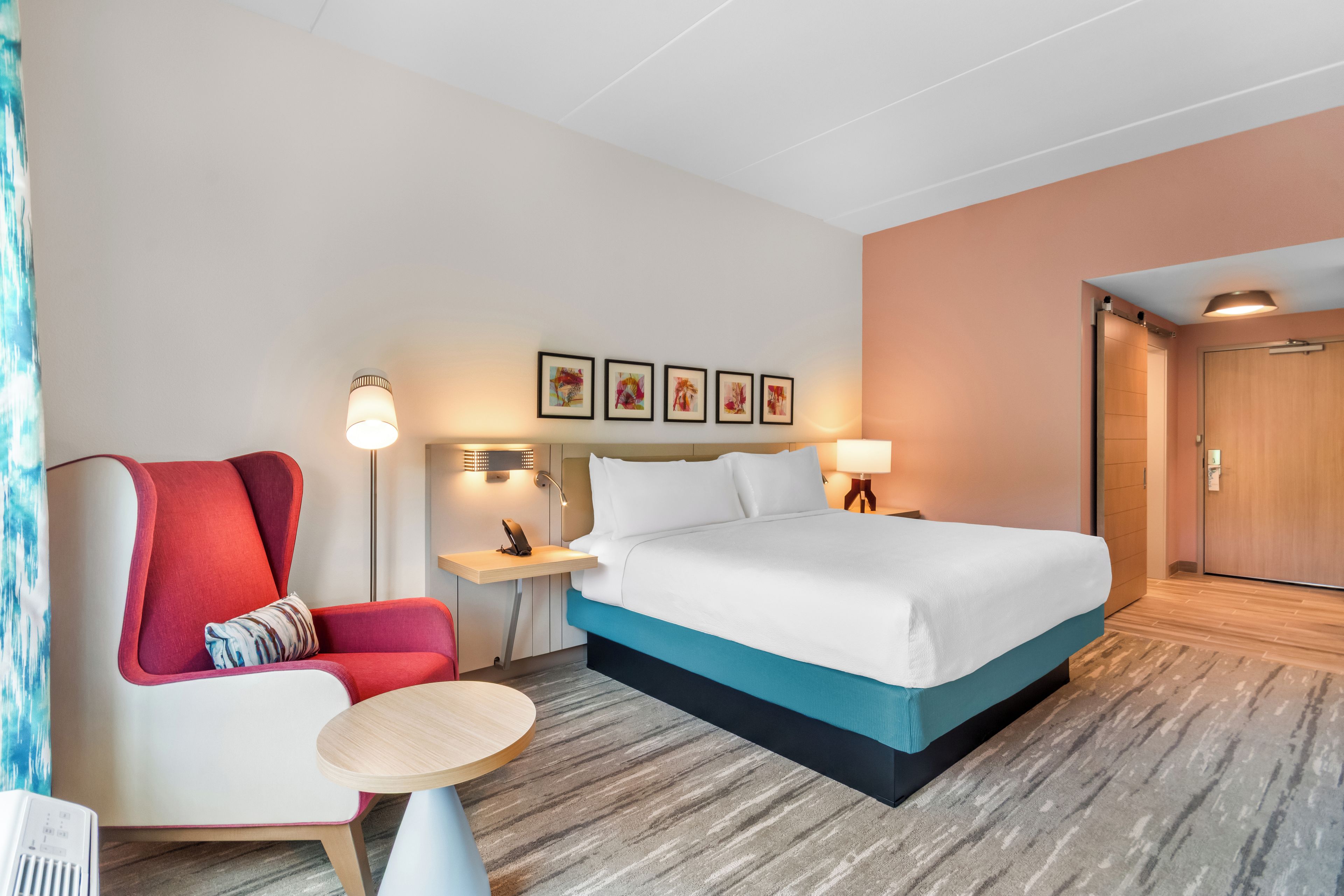 A hotel room with a bed, a chair, a small table, a nightstand, and a wall-mounted lamp.