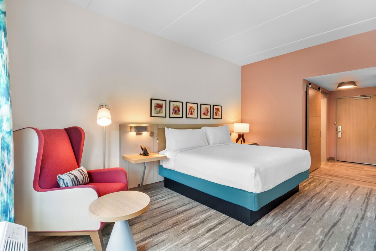 A hotel room with a bed, a chair, a small table, a nightstand, and a wall-mounted lamp.
