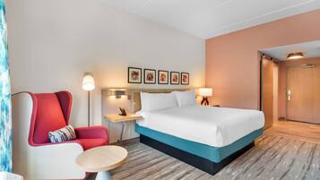 A hotel room with a bed, a chair, a small table, a nightstand, and a wall-mounted lamp.