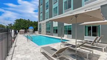 A swimming pool area with lounge chairs and umbrellas, surrounded by a building with large windows and a stone wall.