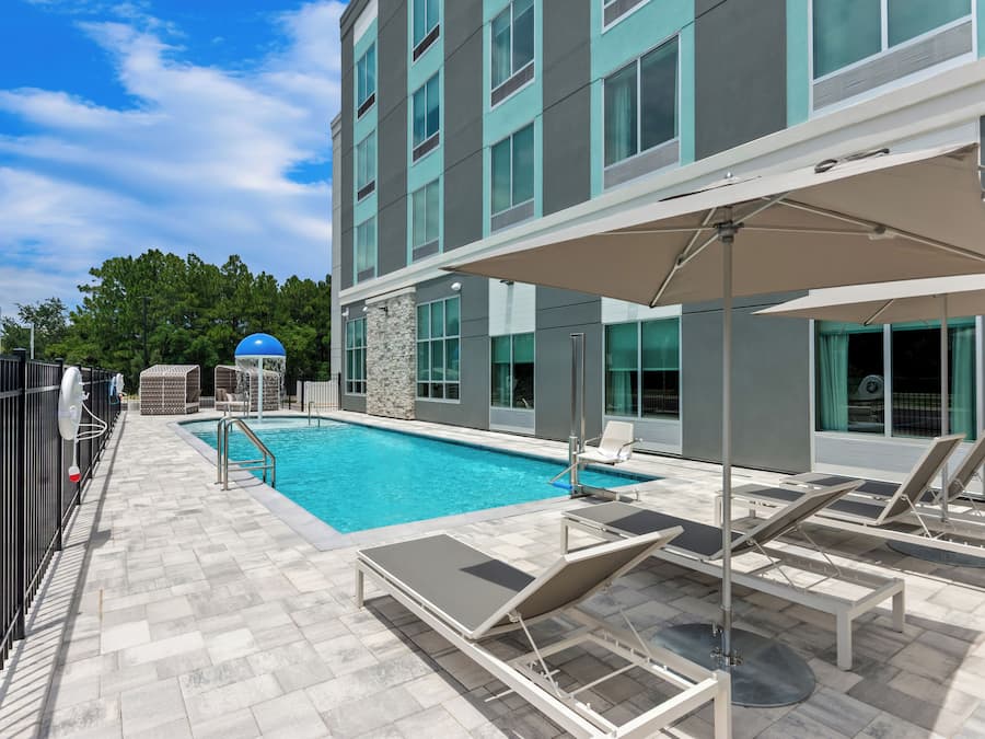A swimming pool area with lounge chairs and umbrellas, surrounded by a building with large windows and a stone wall.