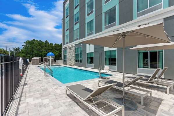 A swimming pool area with lounge chairs and umbrellas, surrounded by a building with large windows and a stone wall.
