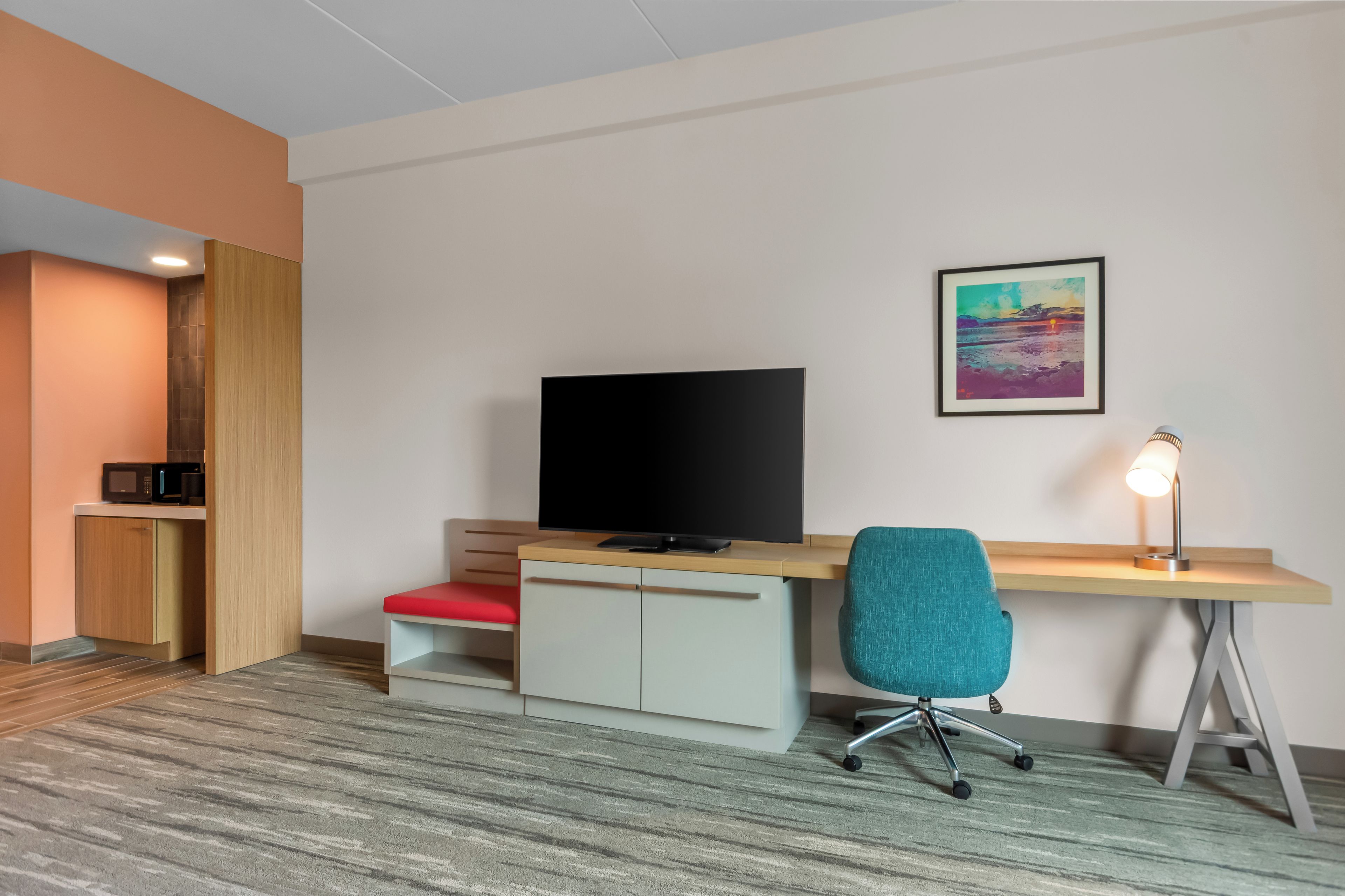 Una habitación de hotel moderna con un televisor de pantalla plana, un escritorio con lámpara, una silla de oficina color turquesa y un cuadro llamativo en la pared.