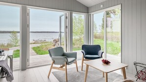 Et moderne værelse med en stor skydedør, der fører ud til en terrasse med udsigt over vandet. Der er to stole og et bord i værelset.