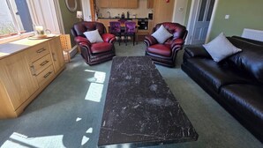 Una sala de estar con dos sillones de cuero, una mesa de centro negra, un mueble de madera y una cocina al fondo.