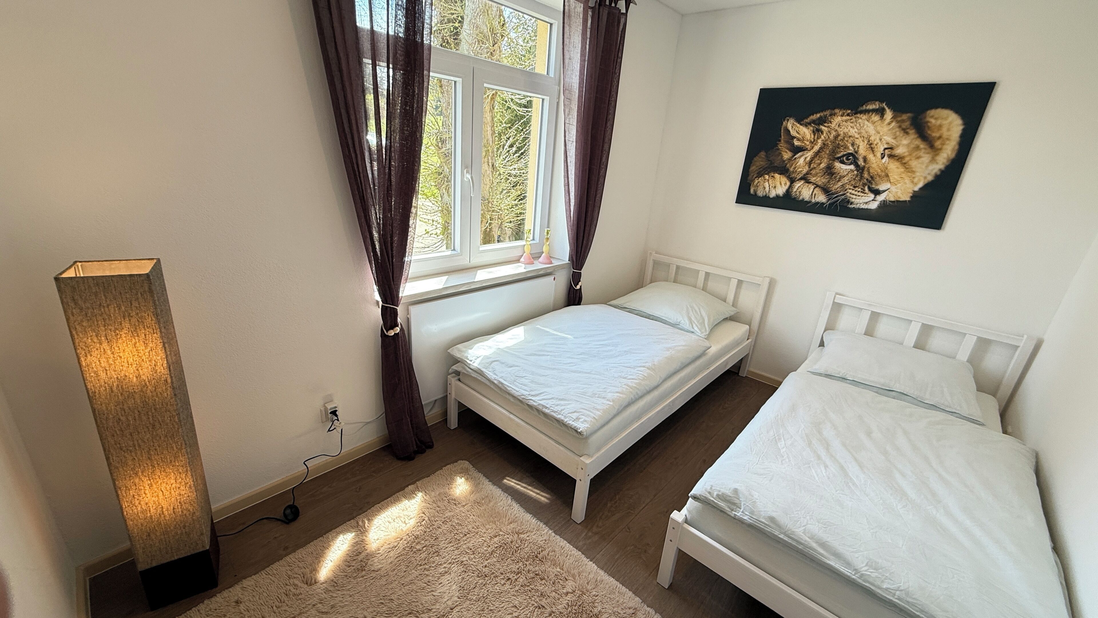 Un dormitorio con dos camas, una ventana con cortinas, un cuadro de un león y una lámpara de pie alta.