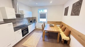 A compact kitchen with a stove, sink, and white cabinets. There's a dining table with chairs and a bench. A window with blinds is visible, and a decorative wall panel is on the right. - Apartments Zum Weihnachtsland "Anton Günther" (Stützengrün)