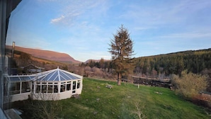 Ein Haus mit Anbauten aus Glas, einer Holzdiele und Blick auf einen Fluss und bewaldete Hügel.
