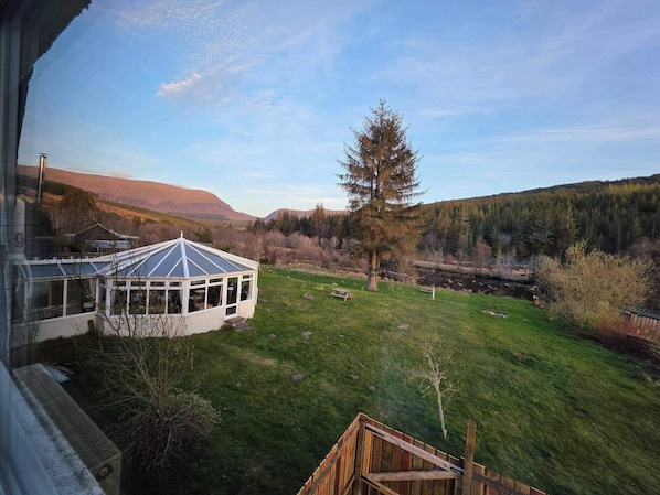 A house with a glass conservatory, a wooden deck, and a view of a river and forested hills.