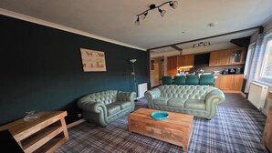 Ein Wohnzimmer mit zwei grünen Ledersofas, einem Holztisch, einem karierten Teppich und einer Küche im Hintergrund.