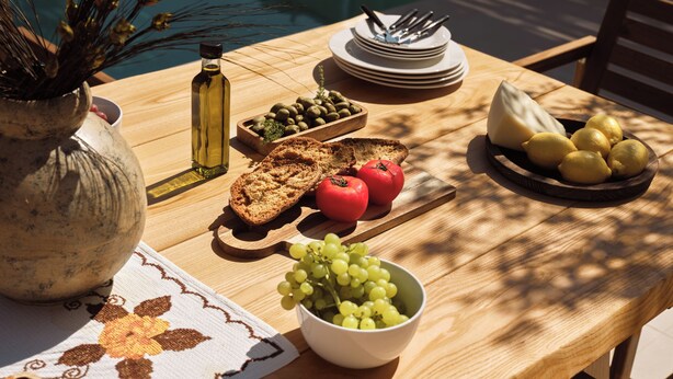 Ein Holztisch mit verschiedenen Speisen, darunter Brot, Tomaten, Trauben, Oliven und ein Käselaib.