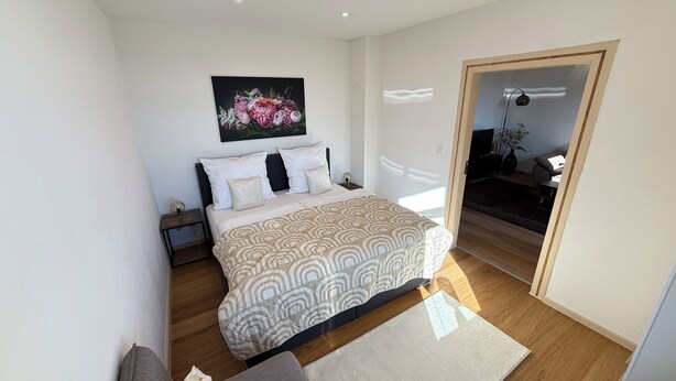 A bedroom with a bed, a nightstand, a wall-mounted painting, and a door leading to another room.
