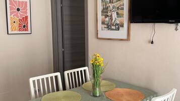Un coin repas avec une table dressée pour quatre personnes, entourée de chaises blanches, d’un tableau encadré et d’un téléviseur fixé au mur.