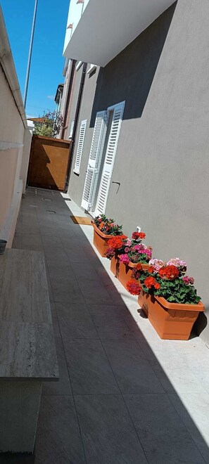 A narrow outdoor corridor with tiled flooring, a row of flower planters, and a building with a grey facade and white shutters. - 50 special apartment (Pontedera)
