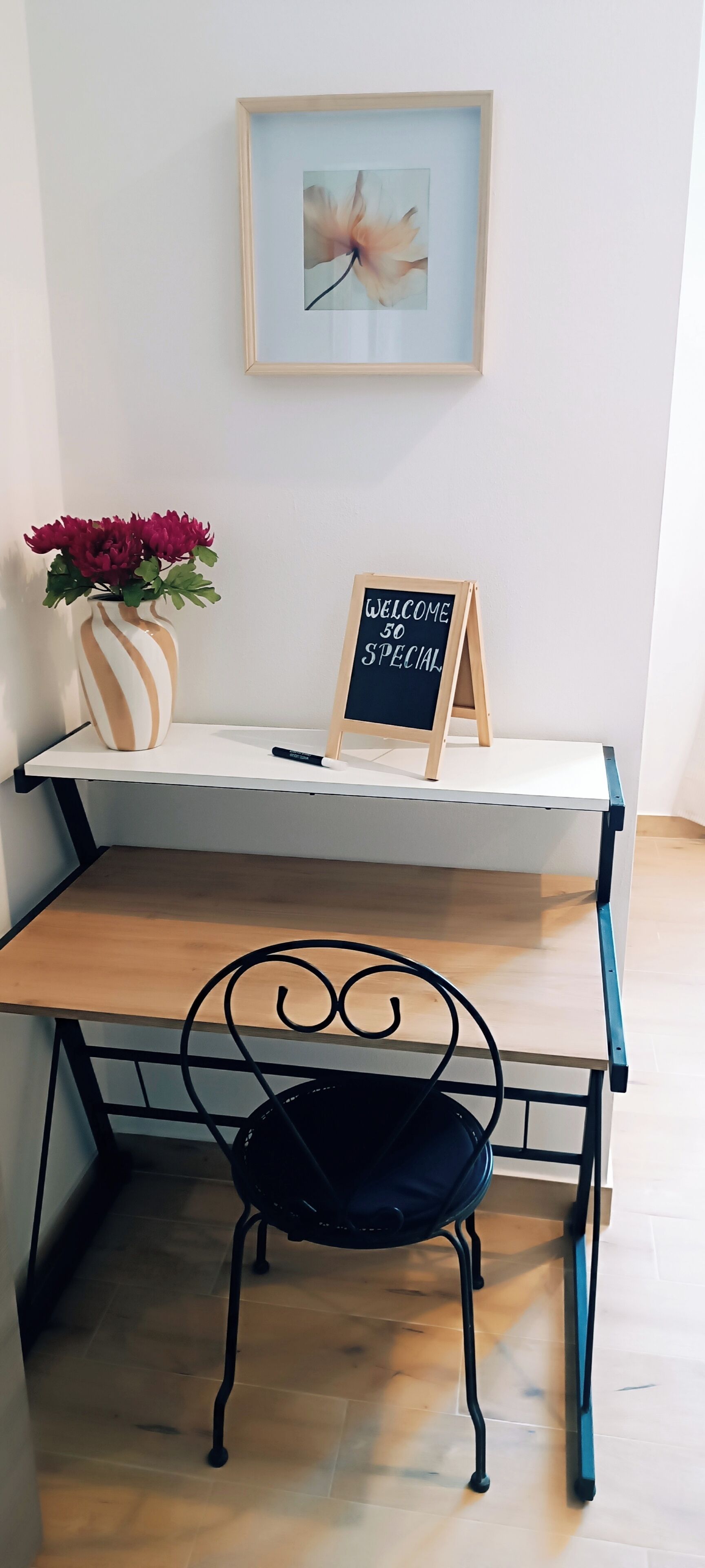Ein Schreibtisch mit Stuhl, ein gerahmtes Bild an der Wand, eine Vase mit Blumen und ein Kreidetafel-Schild.