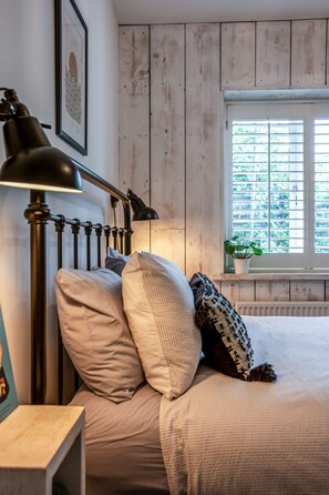 Ein Schlafzimmer mit einem Bett, einem Nachttisch, einer Lampe, einem Fenster mit Jalousien und einer Zimmerpflanze.