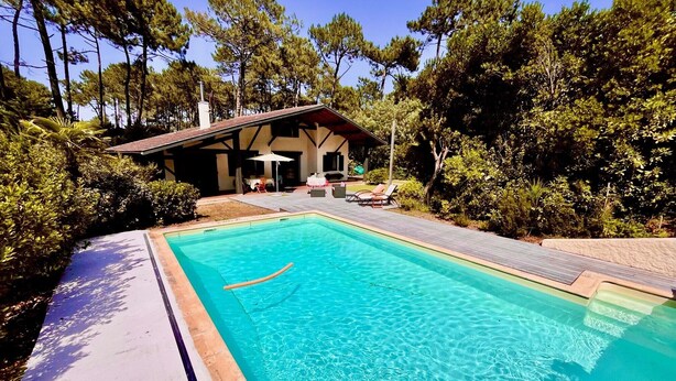 Ein rechteckiger Swimmingpool mit klarem, blauem Wasser, umgeben von einer Holzterrasse. Es gibt einen überdachten Außenbereich mit Sitzgelegenheiten und einem Tisch.
