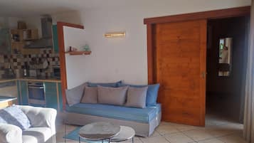 A living room with a sofa, a small round table, a kitchenette, and a wooden door.