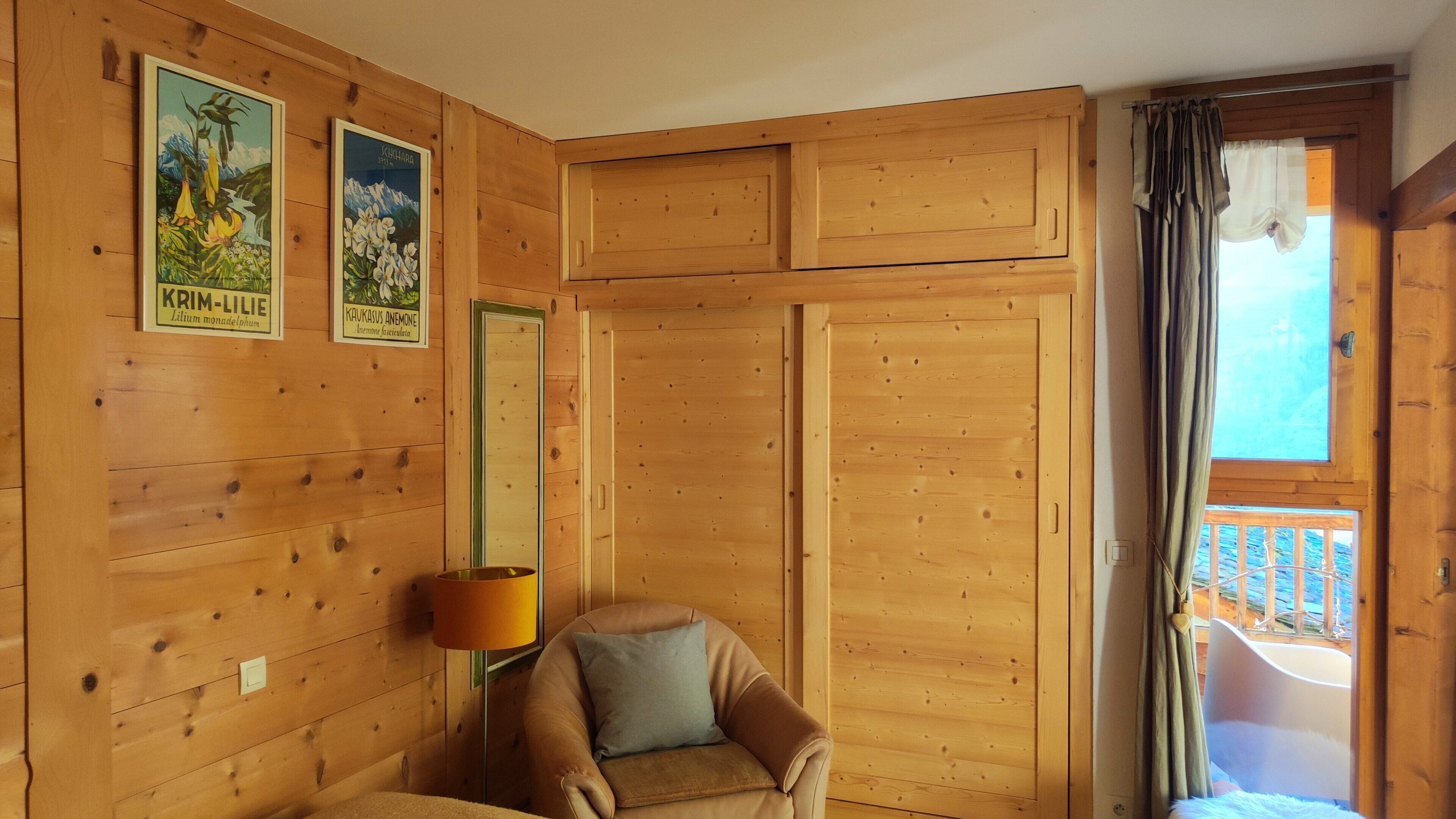 Interior de una cabaña de madera con una sola silla, una lámpara y dos carteles botánicos enmarcados en la pared.
