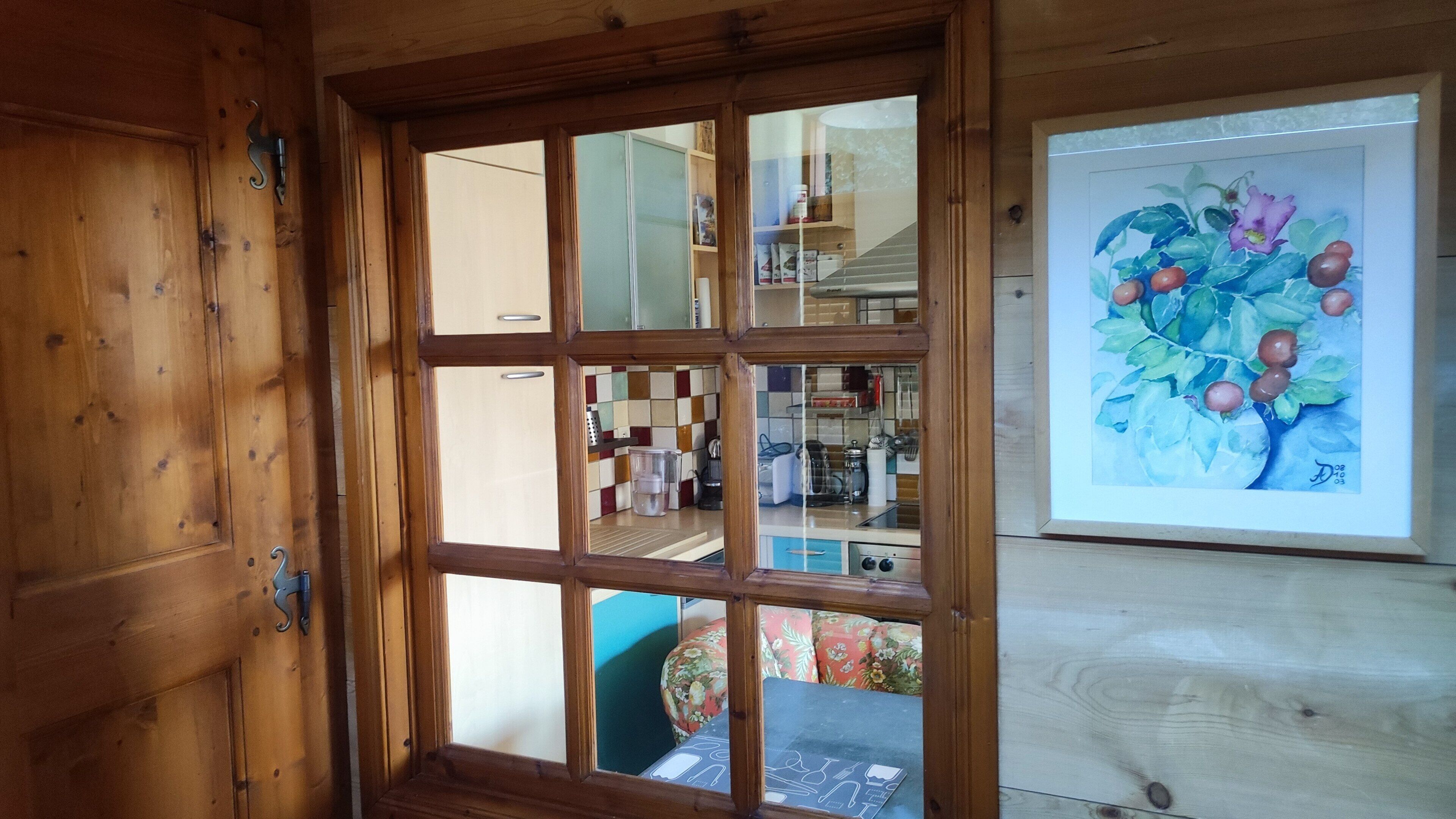 A wooden-framed window with a view into a room containing a kitchen area with cabinets and a painting of fruit on the wall.