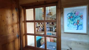 A wooden-framed window with a view into a room containing a kitchen area with cabinets and a painting of fruit on the wall.
