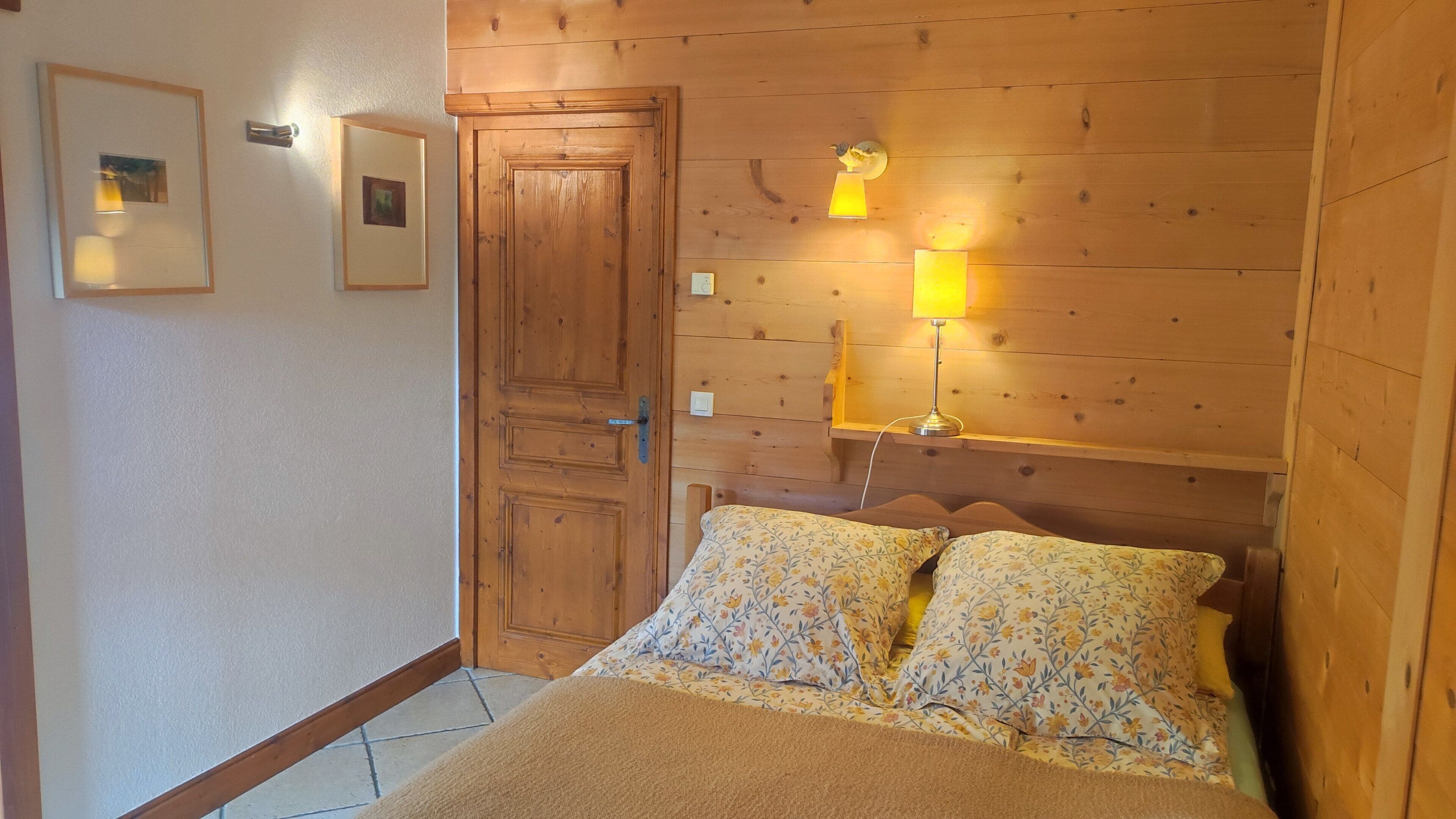 A bedroom with a wooden headboard, a bed with floral bedding, two wall-mounted lamps, and a door.