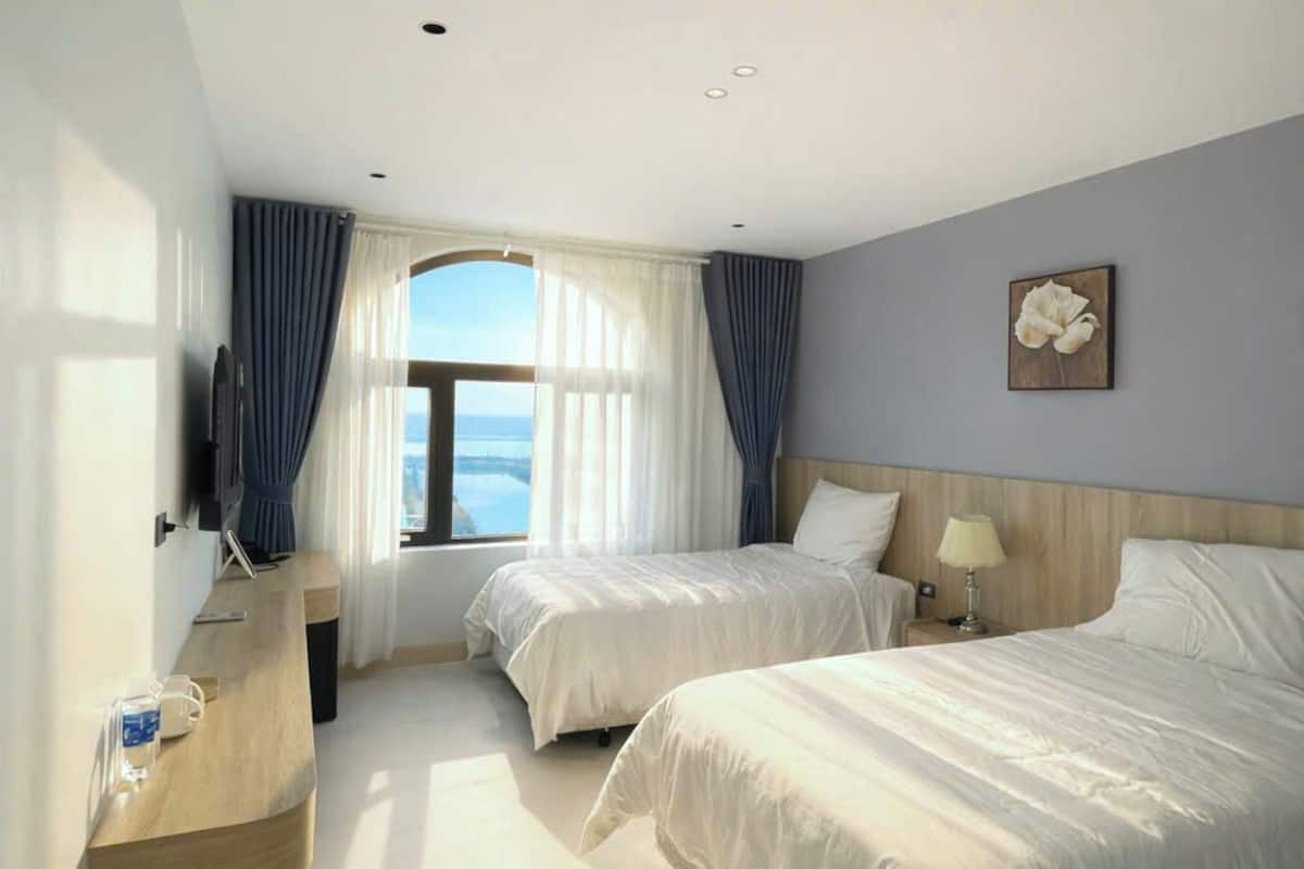 Ein Hotelzimmer mit zwei Betten, einem Fernseher, einem Holztisch, einem Fenster mit Vorhängen und einer Wandblume.