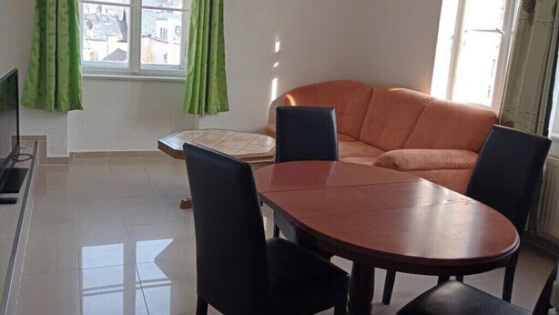 Una habitación con una mesa de comedor, sillas, un sofá y ventanas amplias con cortinas.